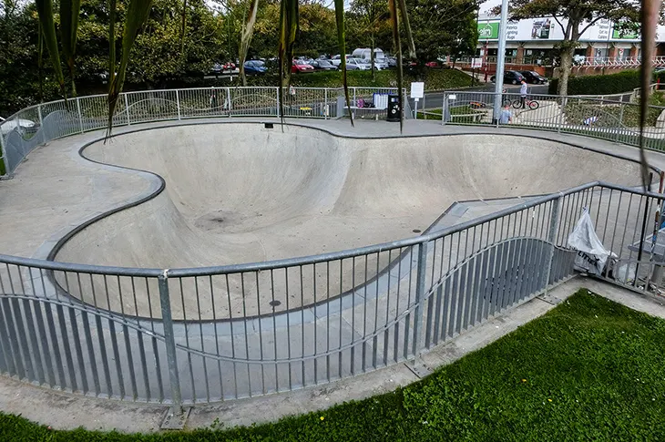 Empty concrete skate bowl surrounded by metal railings and fencing, with trees and parked cars visible beyond.