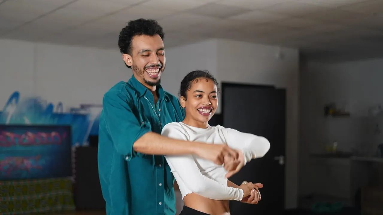 An adult instructor smiles while teaching a young person a dance move in a studio space.