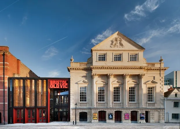Cream-coloured Georgian theatre building with columns and balustrade, flanked by modern red-fronted structure and brick building under blue sky.