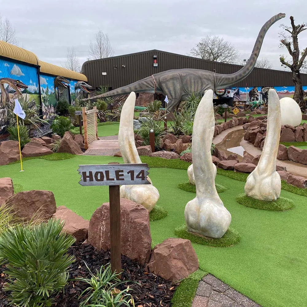 Hole 14 of a dinosaur-themed adventure golf course with a long-necked dinosaur statue, giant egg sculptures, and themed décor.