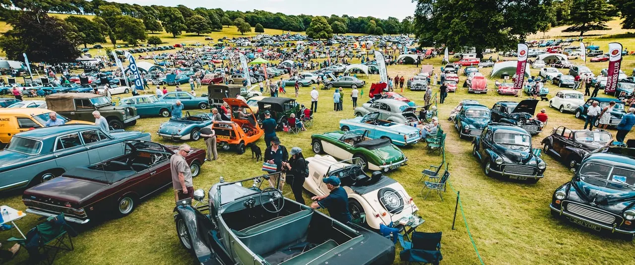 Large outdoor classic car rally with hundreds of vintage vehicles displayed across a grassy field, surrounded by trees and visited by crowds of enthusiasts.