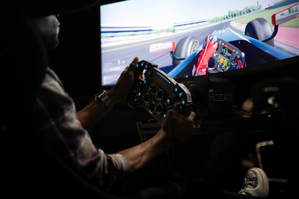 Person holding a racing wheel controller whilst playing a motorsport simulation game on a large screen.
