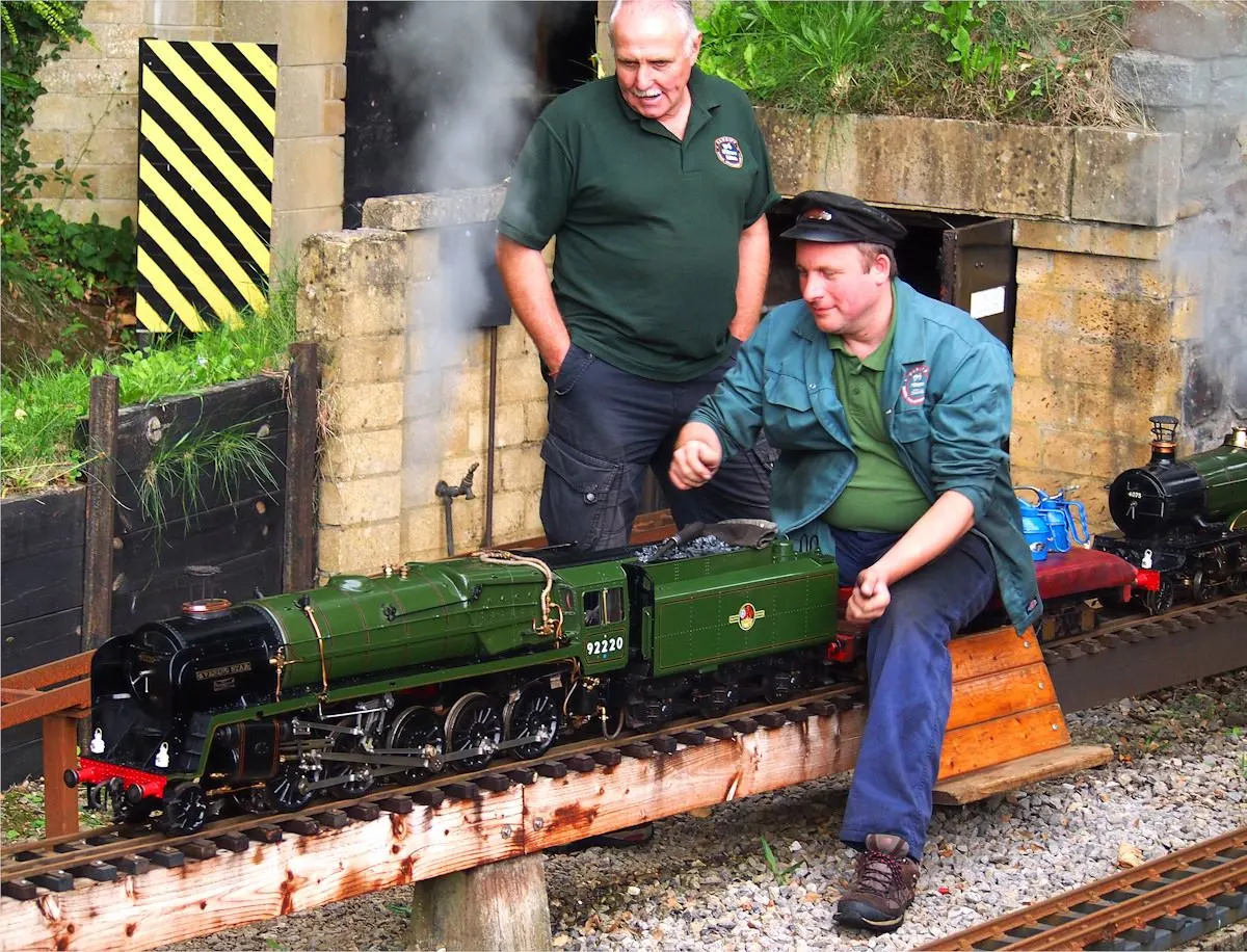 Two staff members operate a miniature steam locomotive on narrow-gauge track at an outdoor railway attraction.