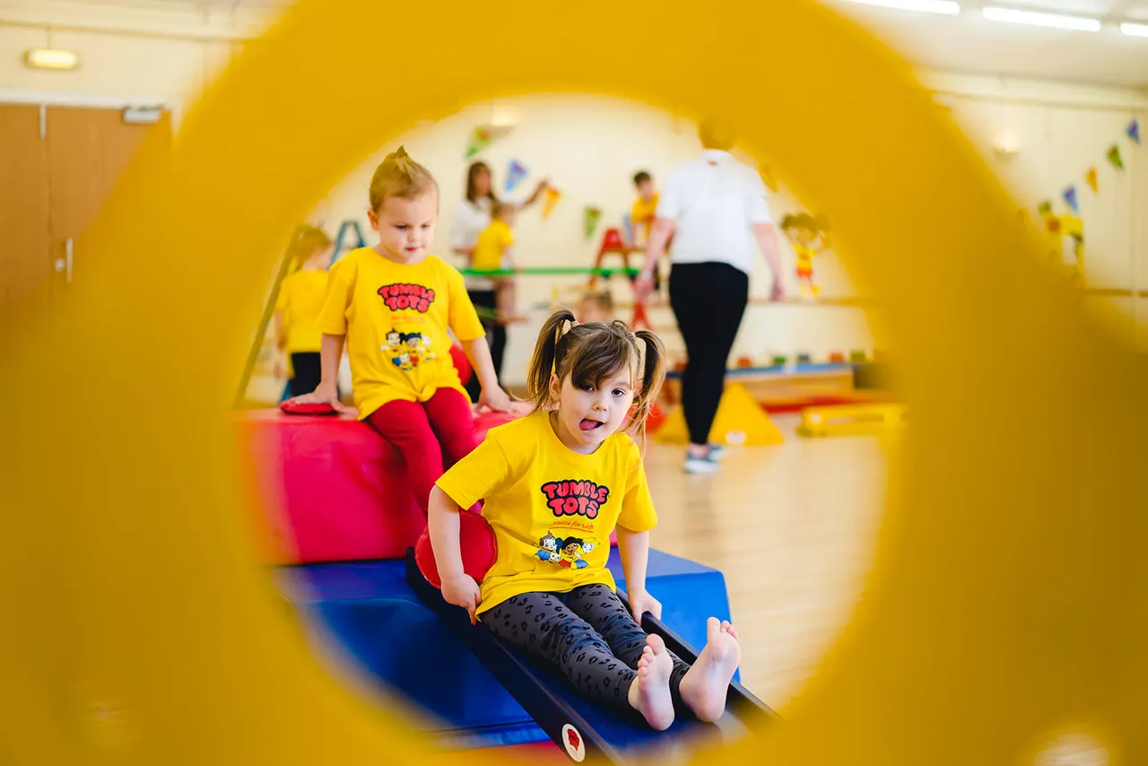 Young children in yellow shirts playing on colourful soft play equipment indoors, viewed through a yellow tunnel.