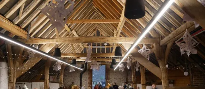 Interior of a rustic wooden barn decorated with white snowflake ornaments and linear lights, with people gathered below.