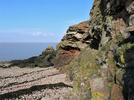 Rocky coastal cliffside with shingle beach and calm sea under clear blue sky.