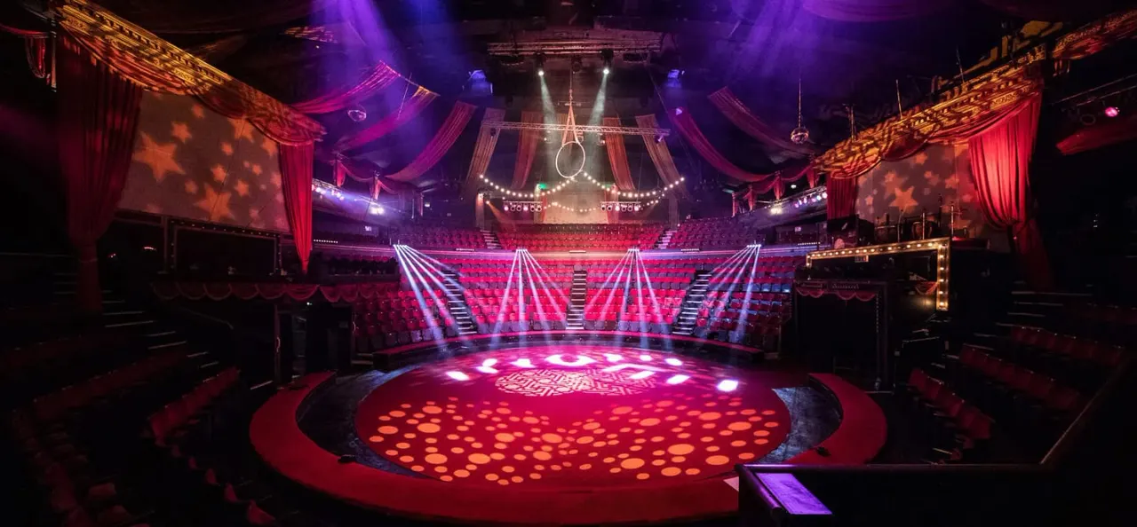 Brightly lit circus ring with red floor and seating, dramatic purple and gold stage lighting, and aerial apparatus suspended above.