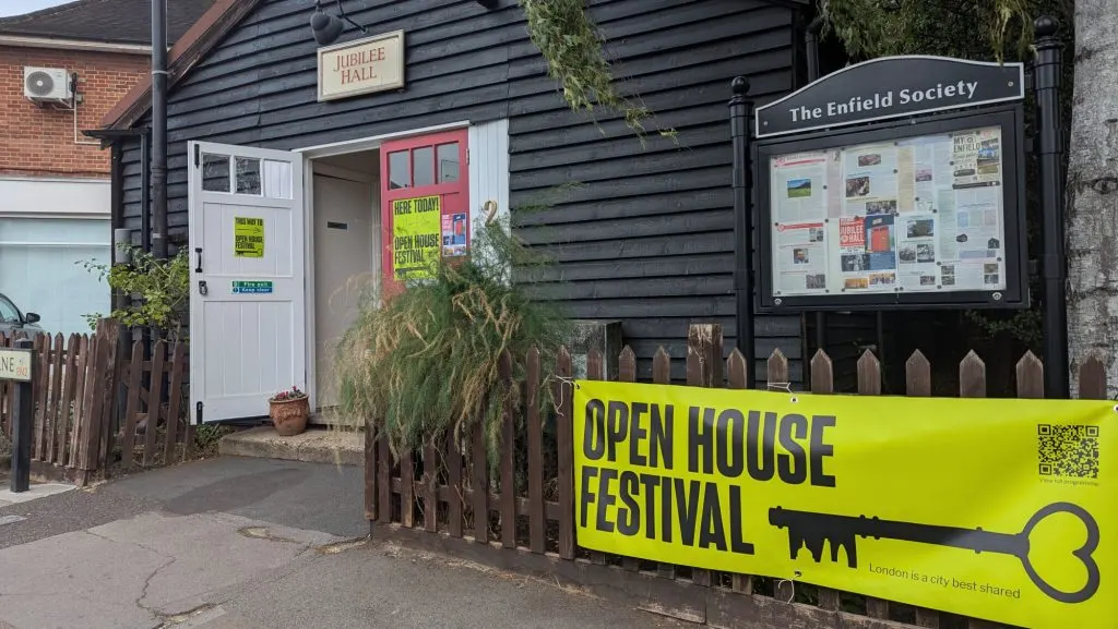 Exterior of a brick and timber-clad community hall with white door, displaying an Open House Festival banner and notice board.