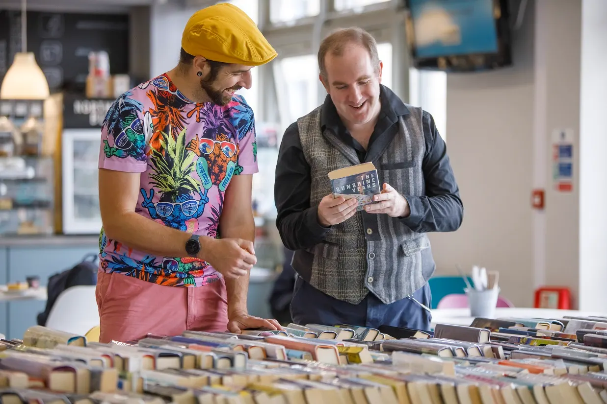 Two men browsing vinyl records together in a library, one wearing a yellow cap and colourful shirt, the other holding an album.