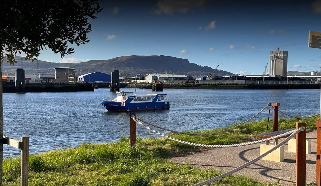 Blue and white passenger ferry moored on calm water with industrial waterfront and mountain backdrop.
