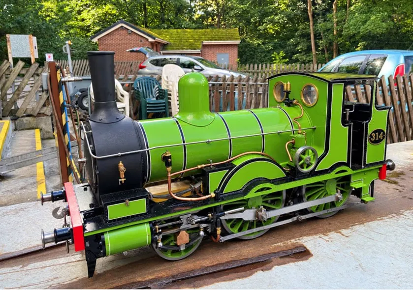 Bright green miniature steam locomotive numbered 314 on display tracks at leisure venue with wooden fences and buildings in background.
