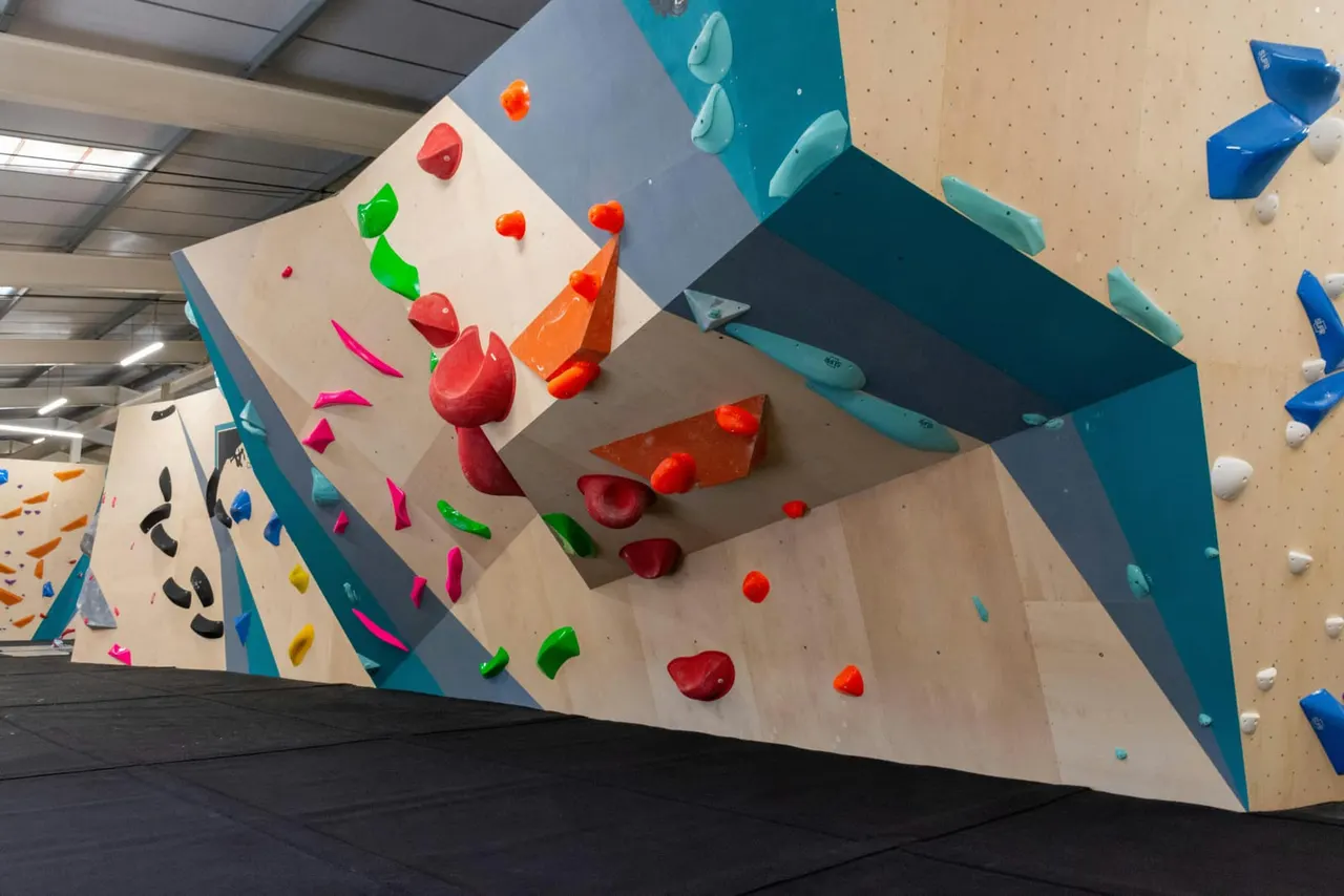 Indoor rock climbing wall with multicoloured handholds of various shapes set into angled panels in a modern sports facility.