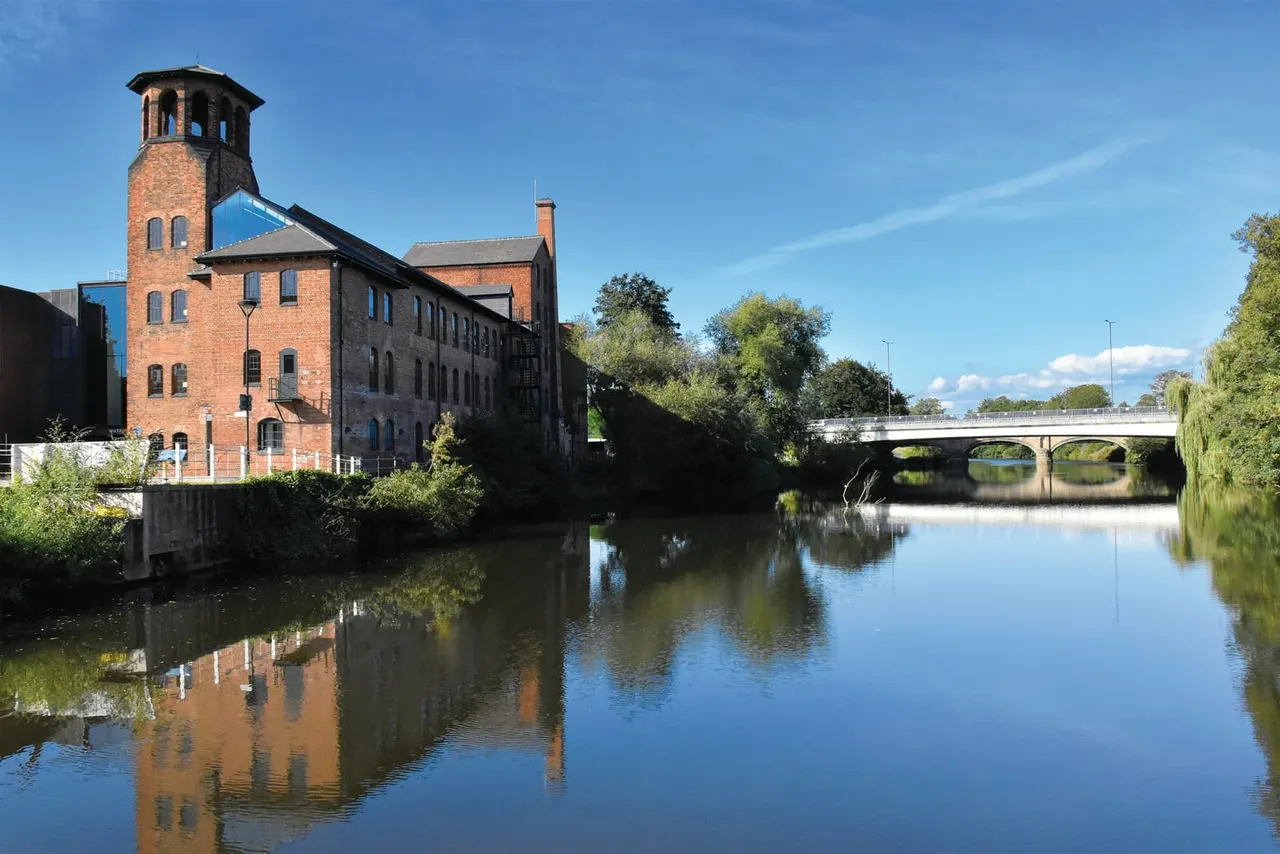 Historic red-brick mill building with tower beside calm river under blue sky, with bridge and trees in background.