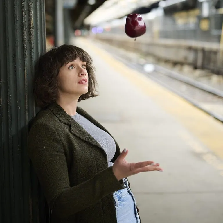 Woman in olive coat tosses a red apple in the air whilst standing on a railway platform.