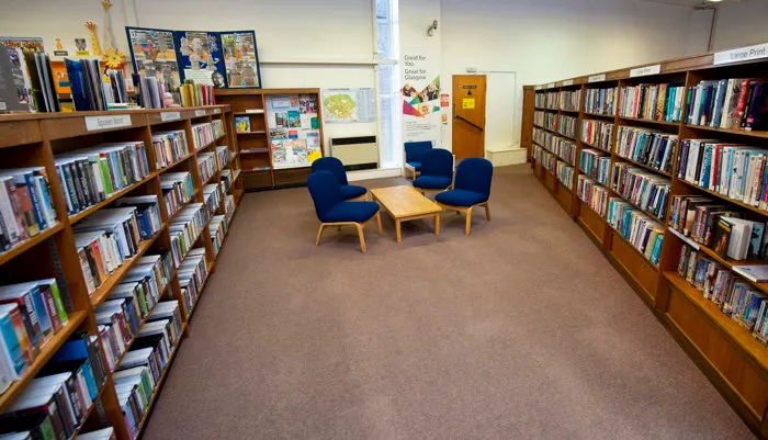 Library interior with wooden bookshelves lining both sides, blue armchairs and wooden table in centre, and various books and educational materials displayed.