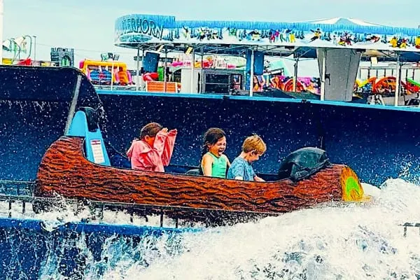 Children playing in a boat-shaped sandbox on a beach with rides visible in the background.