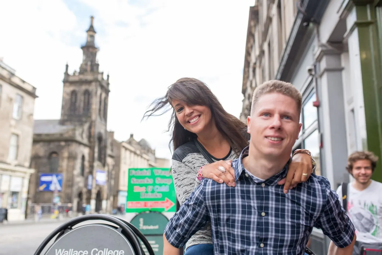 A man gives a woman a piggyback ride on a historic city street with a church tower and stone buildings in the background.