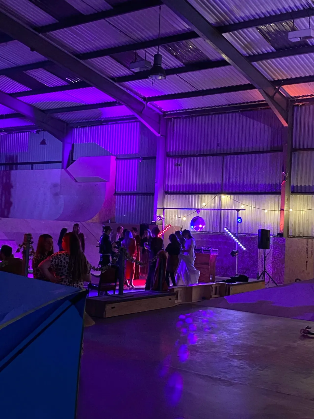 Indoor event space with purple neon lighting, guests mingling on a stage platform, and a dance floor in foreground.