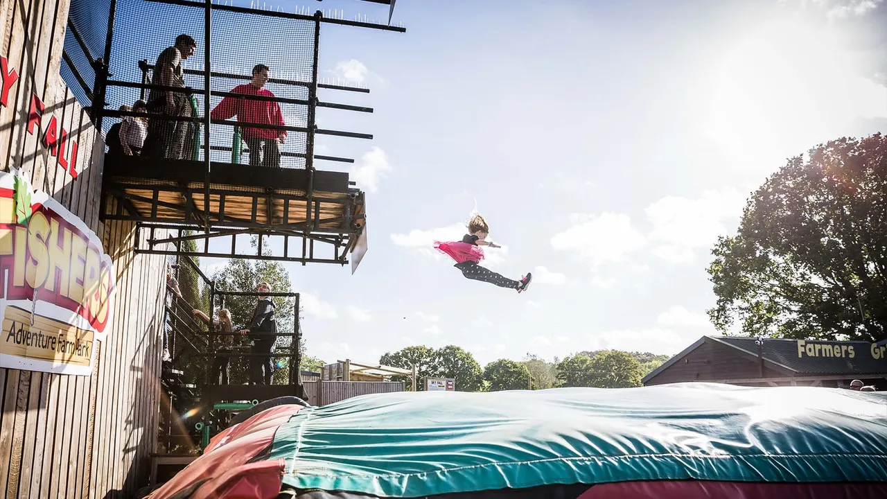 A child in a pink jacket jumps on an inflatable landing bag at an adventure park, with spectators watching from a platform.