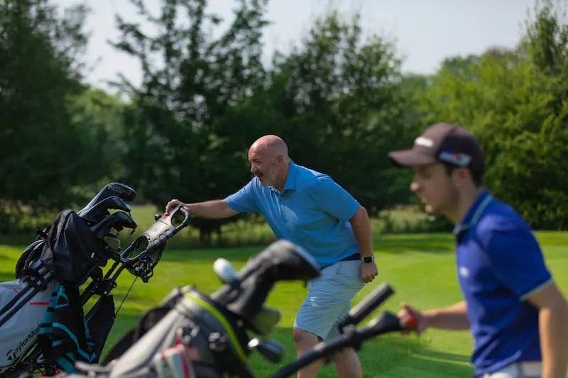 A golf instructor in a blue shirt demonstrates club selection to two golfers on a course surrounded by trees.