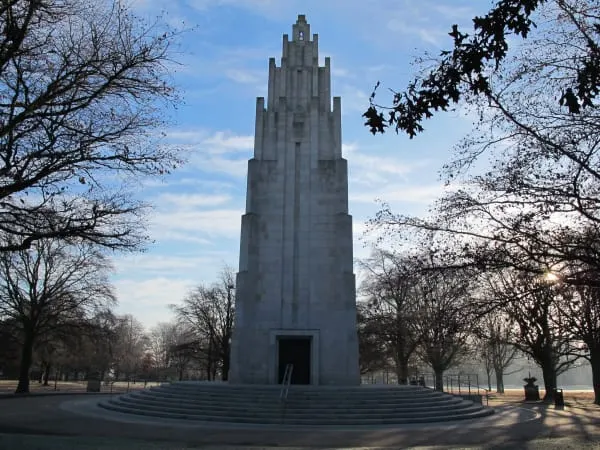 Tall Art Deco war memorial tower surrounded by bare trees in a paved plaza on a clear day.