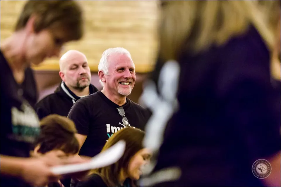 A man smiling whilst holding a small farm animal during an indoor group activity with other participants.