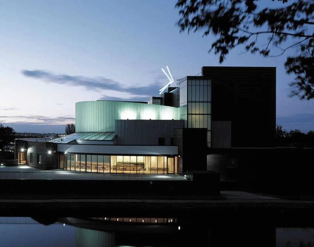 Modern building with green and black metal cladding lit up at dusk, reflected in water below.