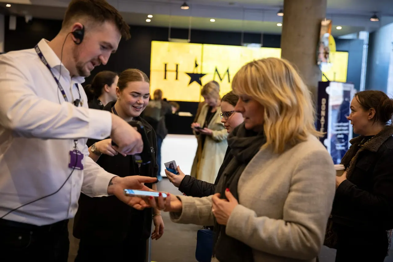 A staff member with a headset serves visitors holding tickets at an indoor venue entrance with a yellow-lit sign.