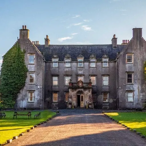 Historic stone mansion with ivy-covered left wing, formal driveway, and manicured lawns under clear sky.