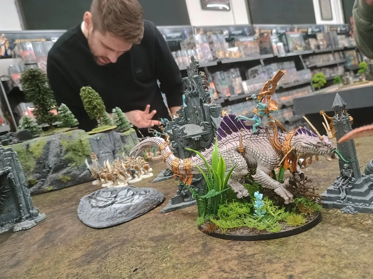 Young person painting a miniature fantasy dragon figure on a gaming table surrounded by castle scenery and terrain pieces.