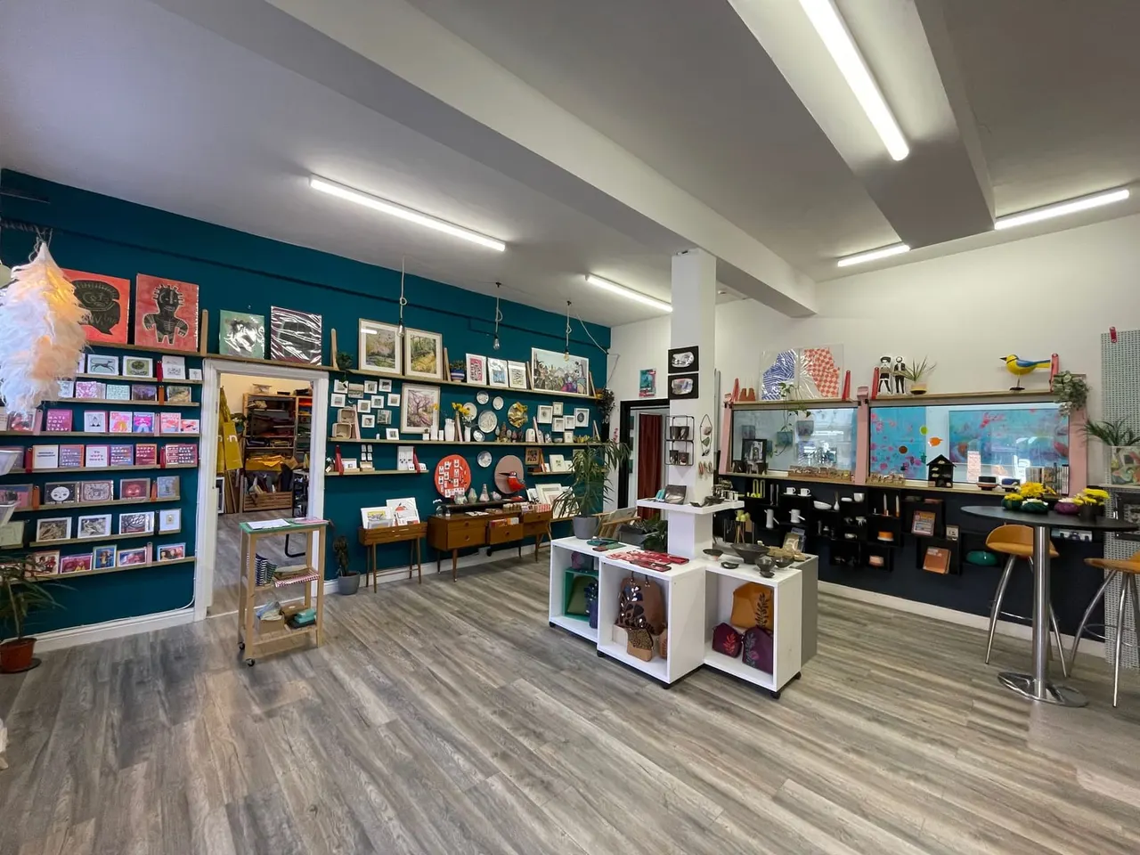 Bright, spacious gallery interior with teal and white walls displaying framed artwork, prints, and decorative objects on shelves and display units.