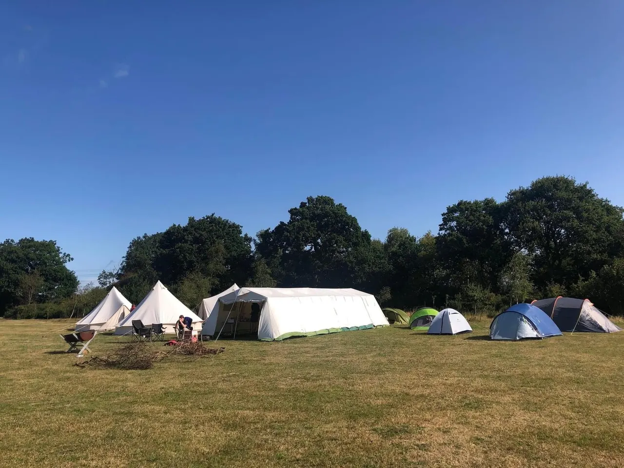 Camping field with various pitched tents, a campfire, and visitors on grassland surrounded by trees under clear blue sky.