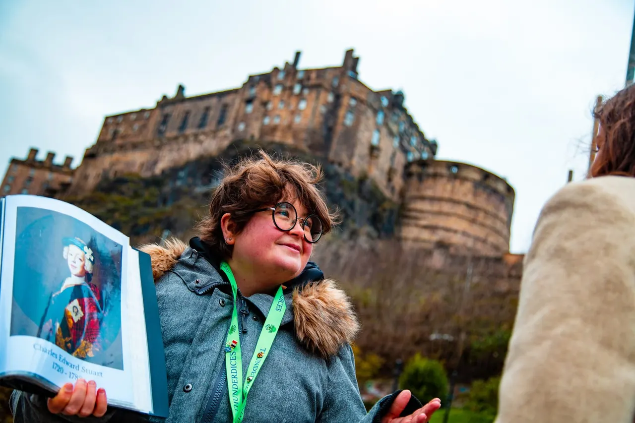 A young visitor wearing glasses and a green lanyard holds an information guide whilst looking up at Edinburgh Castle perched on the rocky hilltop behind.