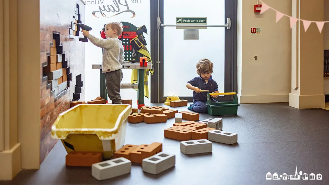 Young children playing with giant building blocks and construction tools in an indoor play area.