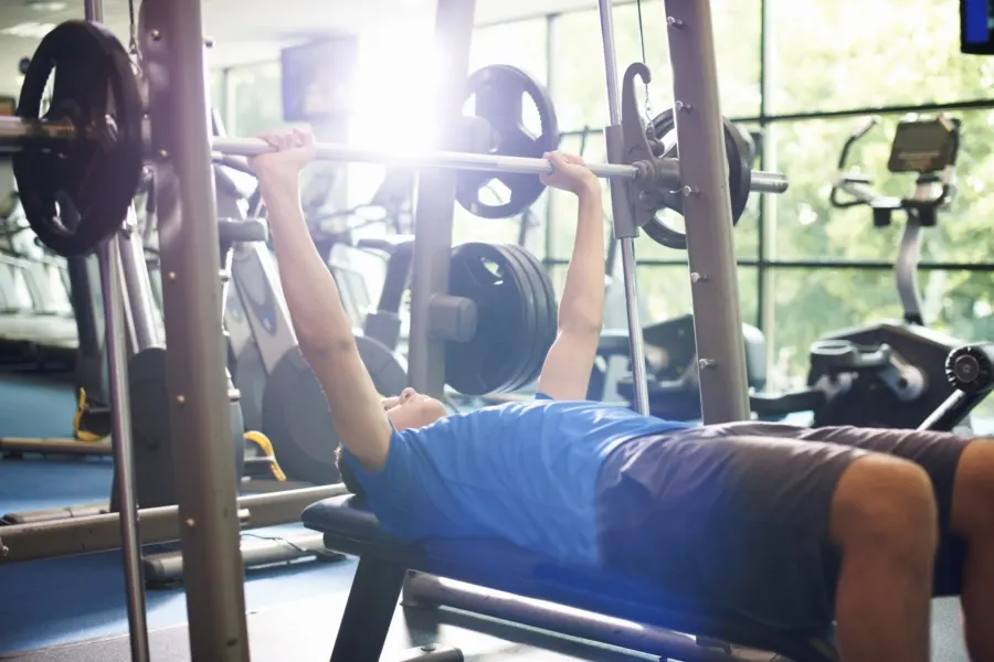Person performing a barbell bench press exercise in a gym with weights and equipment visible.