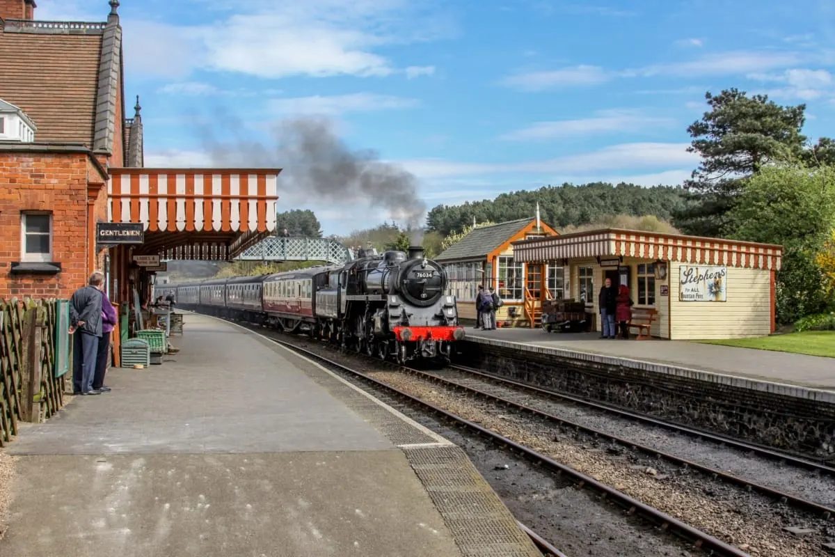 Steam locomotive pulling passenger carriages arrives at heritage railway station with period buildings and visitors on platform.