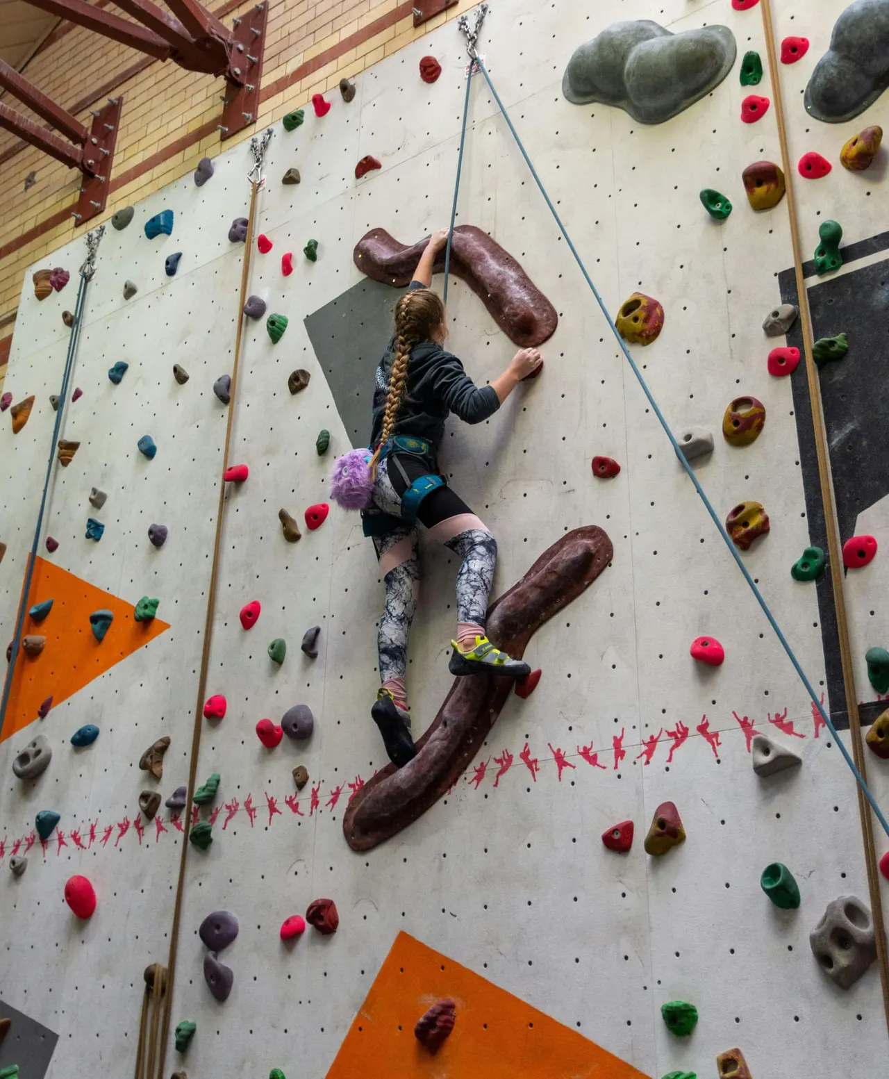 Young person climbing an indoor rock wall with coloured holds, roped in for safety.