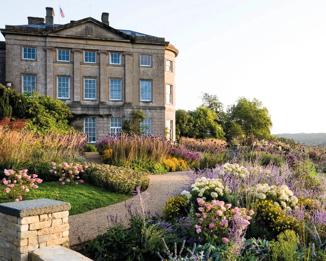 Historic stone mansion with manicured gardens featuring purple and pink flowering plants, lavender borders, and curved pathways.