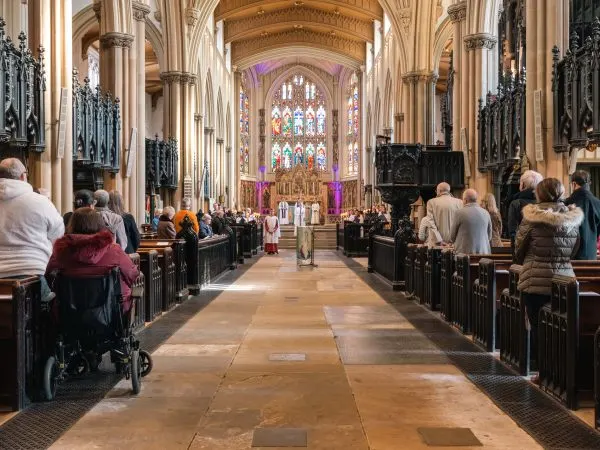 Worshippers and visitors seated in pews inside a grand Gothic cathedral with soaring columns and stained glass windows.