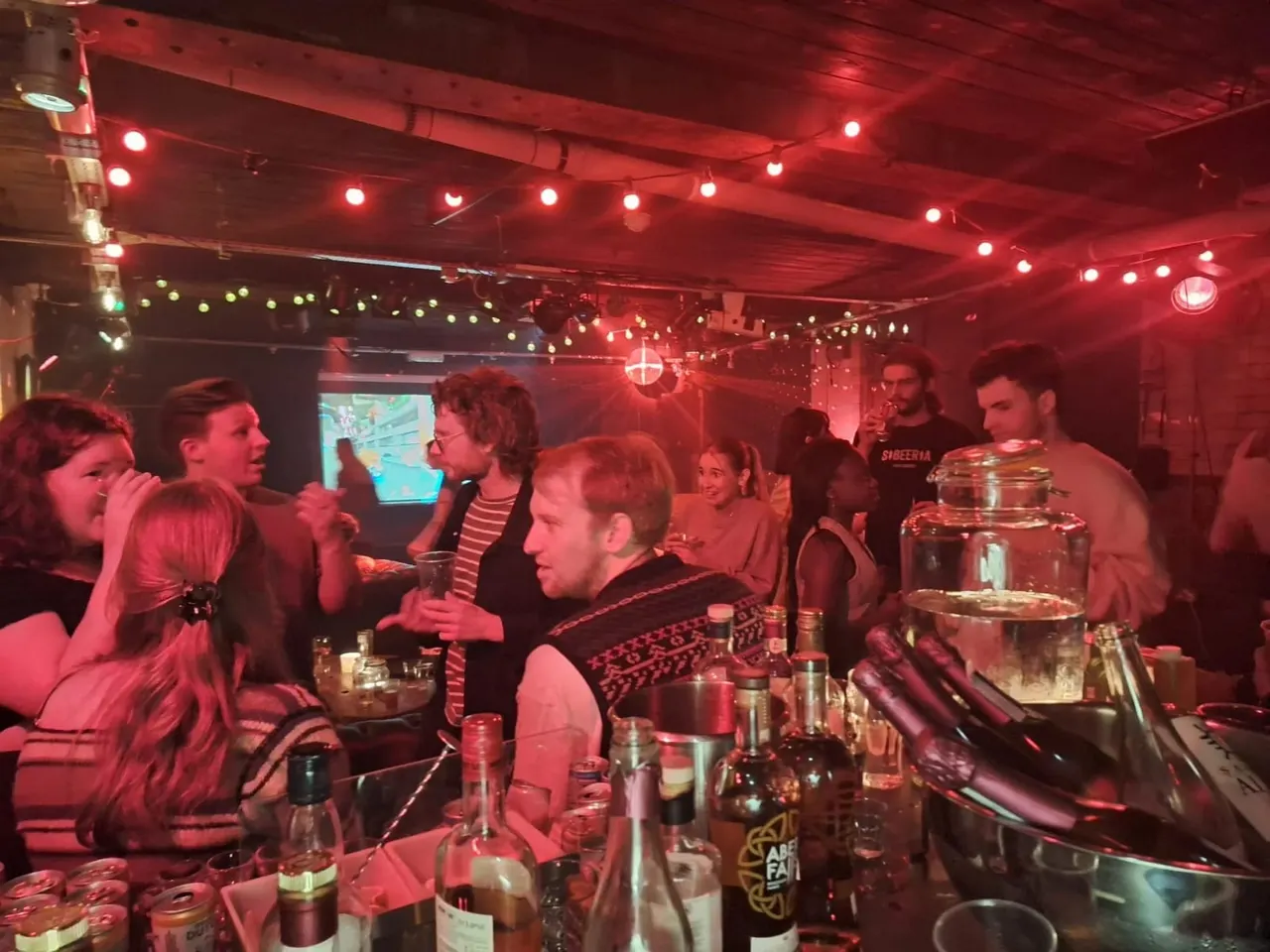 Group of people socialising in a red-lit bar with drinks on the counter and a screen visible in the background.