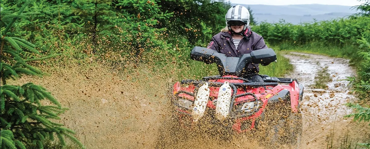 Person riding a red quad bike through muddy terrain, spraying mud, wearing helmet and jacket.