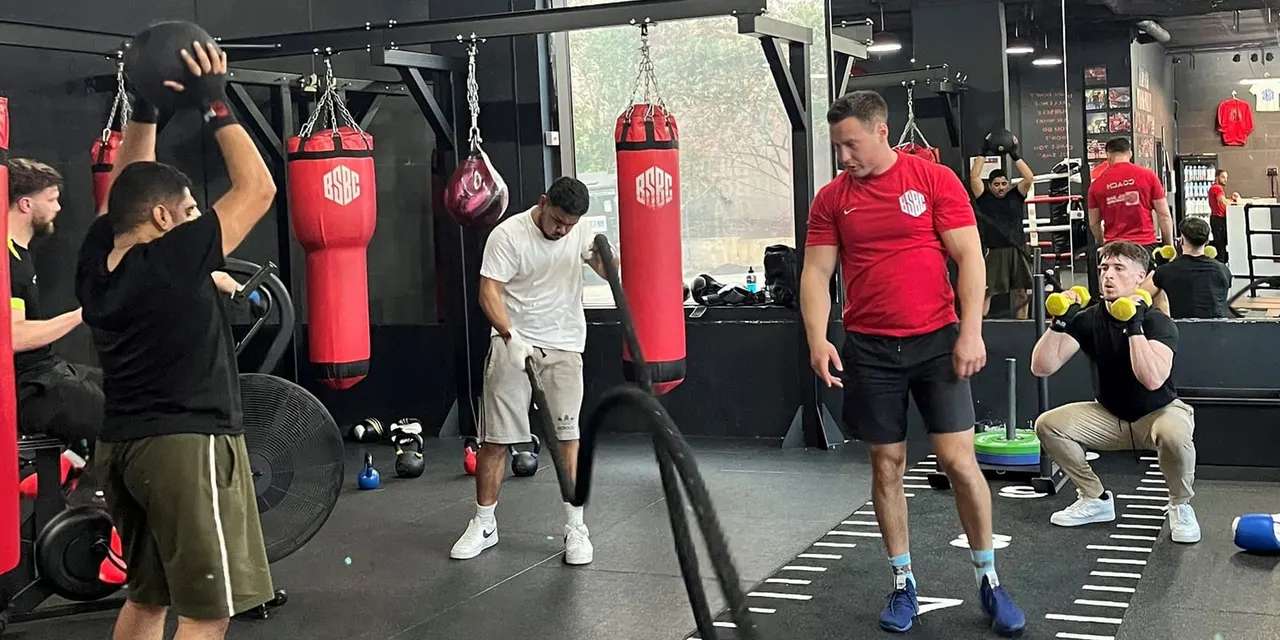 Multiple people training in a boxing gym with heavy bags, weights, and exercise equipment visible throughout the space.