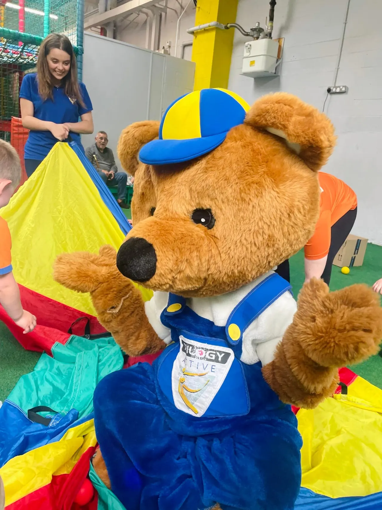 A large brown mascot bear in blue uniform and cap interacts with children playing with coloured parachutes indoors at an active play venue.