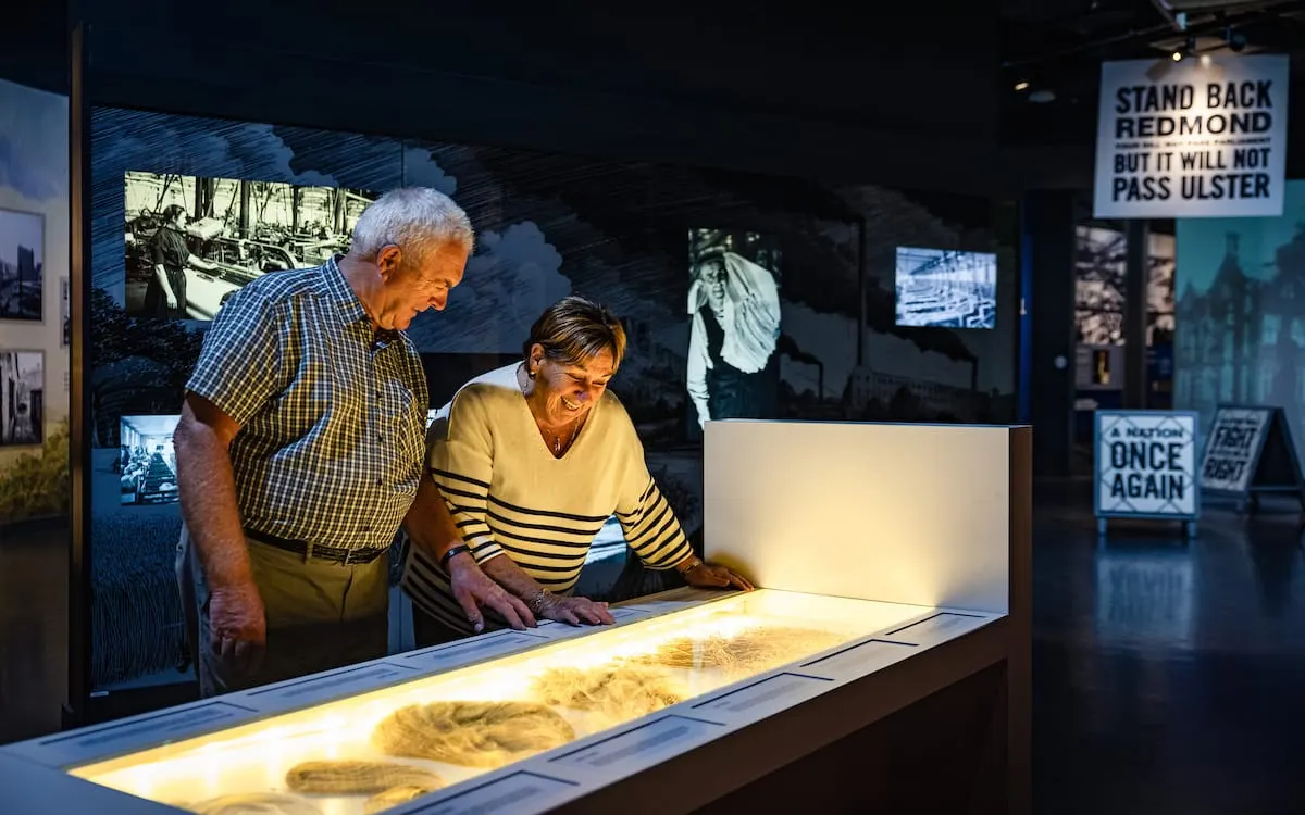 Two older visitors examining an illuminated display exhibit in a museum, surrounded by historical photographs and text panels on the walls.
