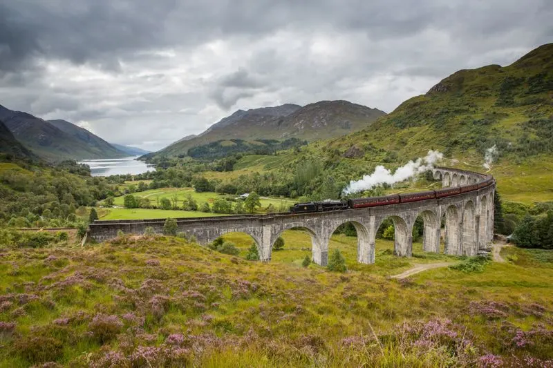 Steam train crossing a stone viaduct through Scottish Highlands with mountains and loch visible.