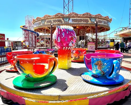 Colourful teacup fairground ride with oversized painted cups in foreground and carousel in background on a sunny day.