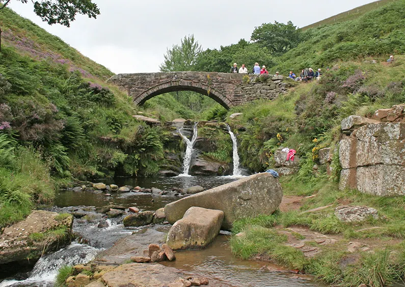 A stream flows through a moorland valley beneath a stone bridge, with visitors scattered across the grassy hillsides.