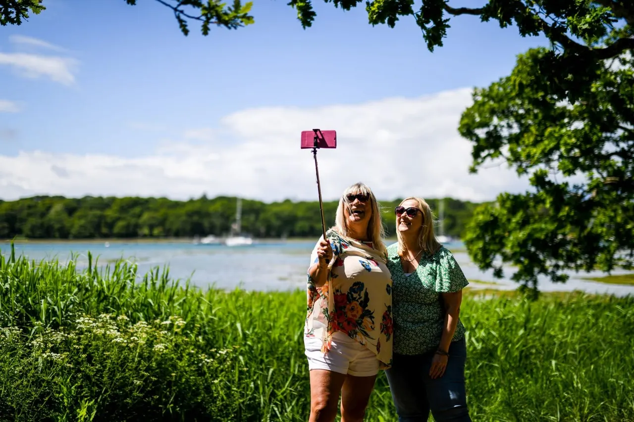 Two women taking a selfie with a pink phone on a stick beside a river, with wooded banks and moored boats visible behind them.