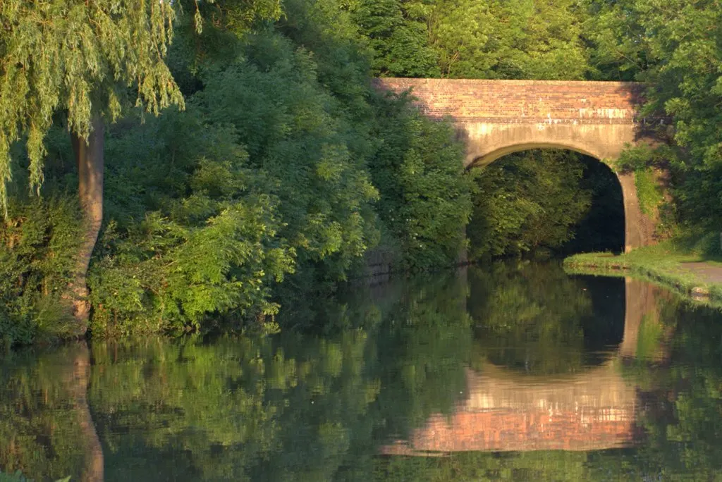 A brick canal bridge surrounded by lush green vegetation with still water reflecting trees and the bridge below.