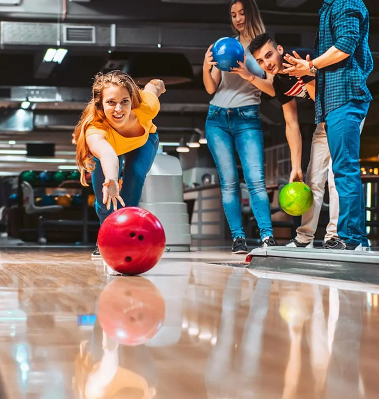 A young woman in a yellow shirt bowls a red ball down a lane whilst friends watch and hold blue and green bowling balls behind her.